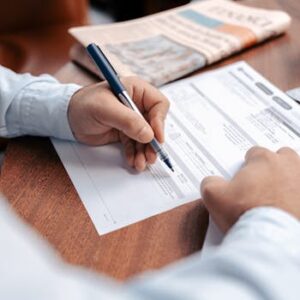 Person reviewing and writing on financial documents with a pen on a wooden desk.