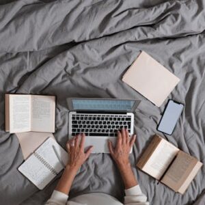 Person typing on laptop surrounded by books and smartphone on a bed, top view.